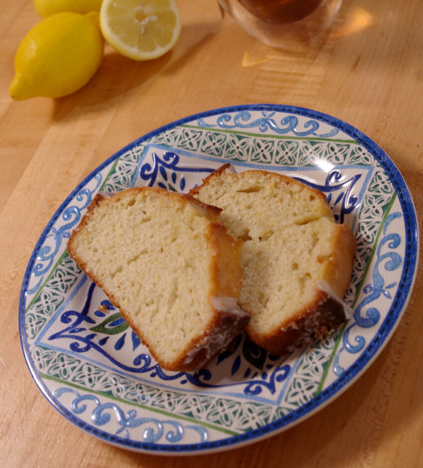 Two fluffy slices of loaf cake with white glaze sit on a patterned blue and white plate on a blond wood counter with cut lemons in the background