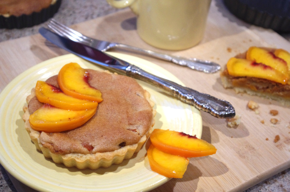 A tart nearly overflowing with brown-colored frangipane is topped with peach slices and arranged on a pale yellow plate with an ornate knife