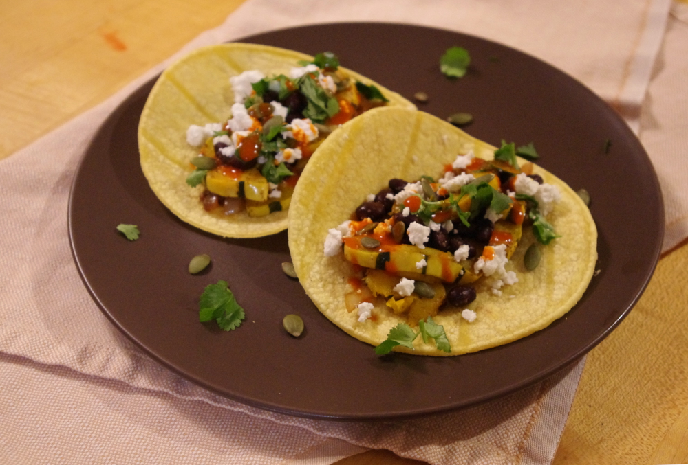 two tacos on a brown plate atop a napkin. The tacos are filled with squash, beans, white cheese, herbs, and a bright drizzle of hot sauce.