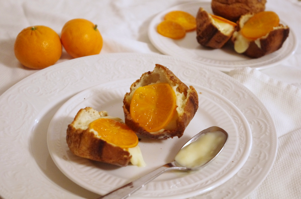 on two ornate white plates, a popover, split in half, topped with cream and clementine slices. in the background another plate, out of focus, and whole clementines