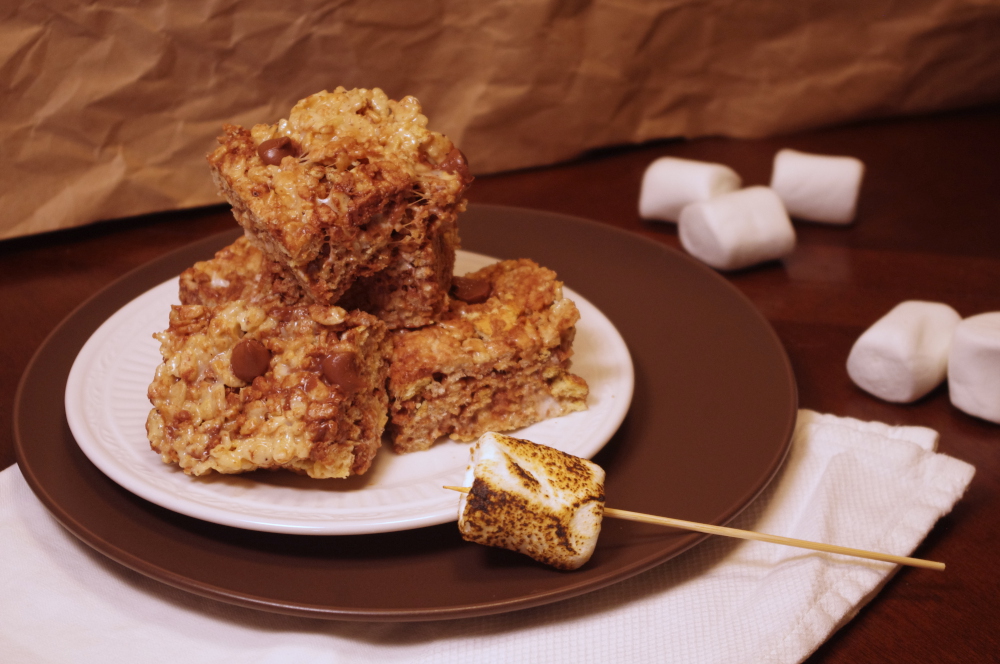 chocolate-studded rice crispy treats stacked on white and brown plates with a toasted marshmallow in the foreground