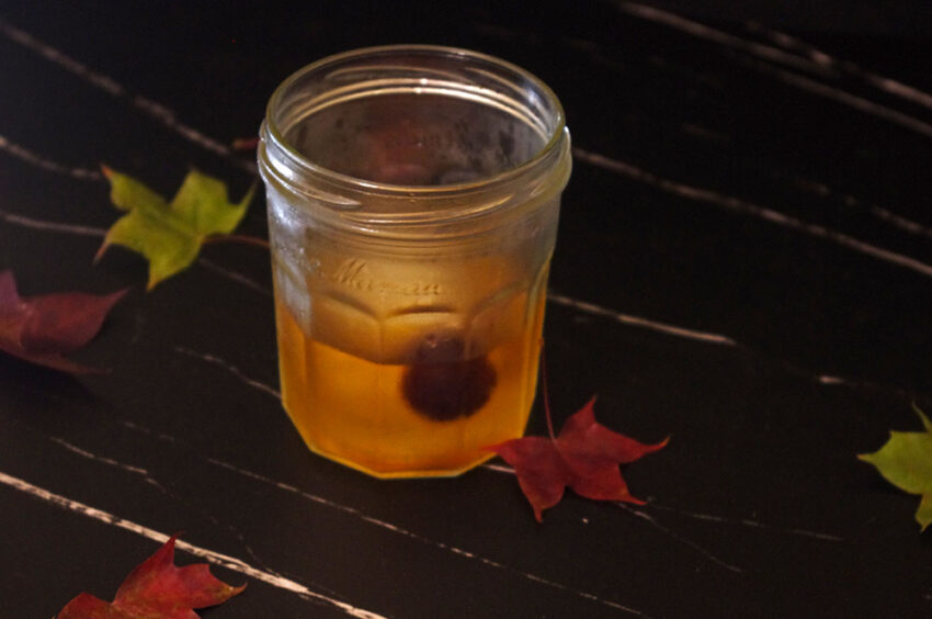 an amber colored brown butter old fashioned with a cherry served in a glass jar surrounded by maple leaves against black marble