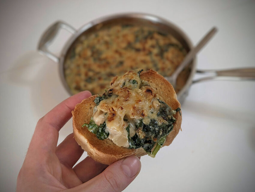 A hand holding a piece of toasted bread with cheesy spinach artichoke beans on top